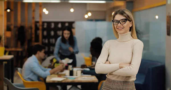 Ofis çalışma alanında duran kameraya bakan genç başarılı iş kadını portresi. Bulanık arka plan üzerinde çalışan insanlar.