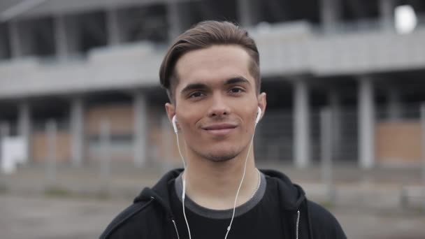 Retrato de corredor masculino atractivo con auriculares que se ejecutan en el fondo del estadio. El atleta mira a la cámara, sonríe. Aptitud saludable atlético hombre corriendo al aire libre . — Vídeo de stock