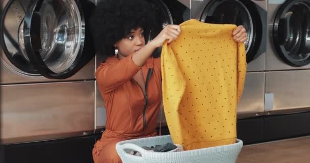 Jovem afro-americana classificando roupa na cesta classificando roupas antes de lavar sentado na lavanderia pública self-service . — Vídeo de Stock