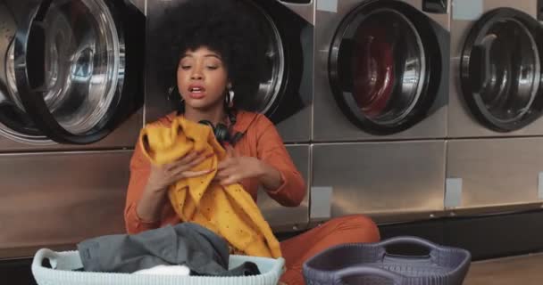 Счастливая афроамериканская молодая женщина сортирует бельё в корзине, сортируя одежду перед стиркой, сидя в общественном стирке . — стоковое видео