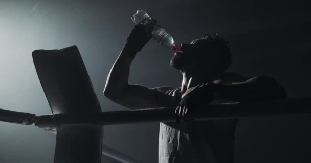 Joven hombre cansado bebiendo de la botella de agua después de entrenar en el anillo de boxeo oscuro con humo. Silueta. Concepto de boxeo . — Vídeos de Stock