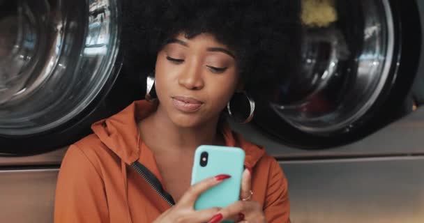 Jeune femme souriante utilisant un smartphone à la laverie automatique. Laverie publique en libre-service . — Video