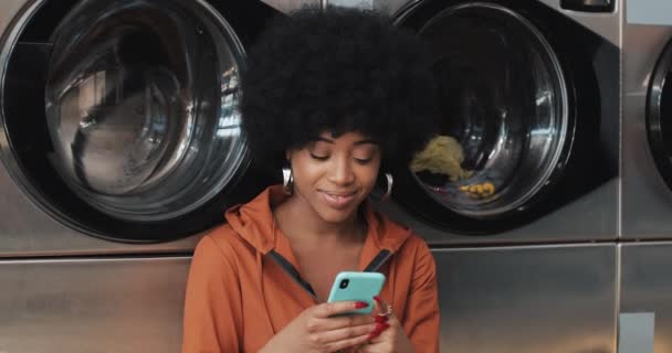 Jeune femme souriante utilisant un smartphone à la laverie automatique. Laverie publique en libre-service . — Video