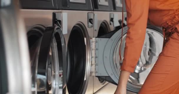 Junge afrikanisch-amerikanische Frau sitzt vor einer Waschmaschine und lädt die Waschmaschine mit schmutziger Wäsche. Öffentliche Wäscherei zur Selbstbedienung. — Stockvideo