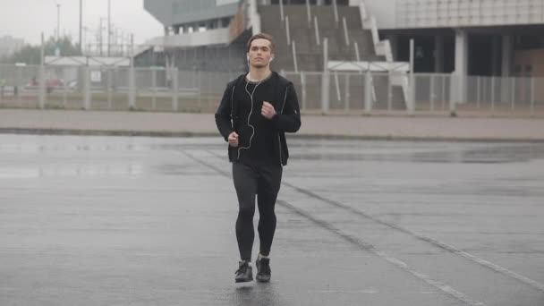Attraktiv ung man kör utomhus bär hörlurar slow motion. Regnigt väder. Konditionsträning träning. Hälsosam livsstil-löpning. — Stockvideo