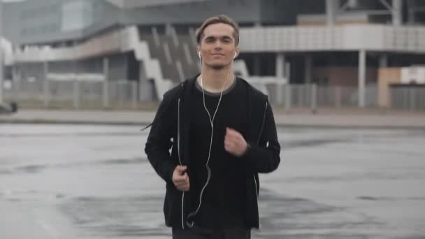 Atractivo joven corriendo al aire libre con auriculares en cámara lenta. Está mirando a la cámara. Clima lluvioso. Entrenamiento de ejercicio cardiovascular. Estilo de vida saludable - correr . — Vídeos de Stock