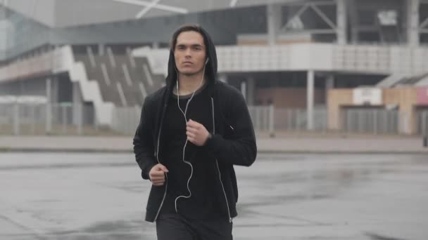 Jovem atraente correndo ao ar livre usando fones de ouvido câmera lenta. Tempo chuvoso. Exercício cardiológico. Estilo de vida saudável - execução . — Vídeo de Stock