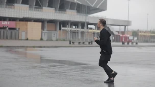 Jovem atraente correndo ao ar livre usando fones de ouvido câmera lenta. Tempo chuvoso. Exercício cardiológico. Estilo de vida saudável - execução . — Vídeo de Stock