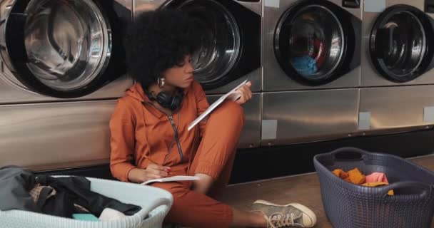 Jovem mulher afro-americana atraente lendo um livro e escrevendo nota enquanto lavava sua roupa na lavanderia . — Vídeo de Stock