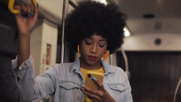 有吸引力的年轻非洲裔美国妇女拿着扶手,在公共交通工具上使用智能手机。夜间时间. — 图库视频影像