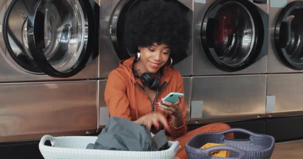 Jovem afro-americana usando smartphone e classificando a roupa na cesta classificando roupas antes de lavar. Ela sentada na lavanderia pública self-service . — Vídeo de Stock