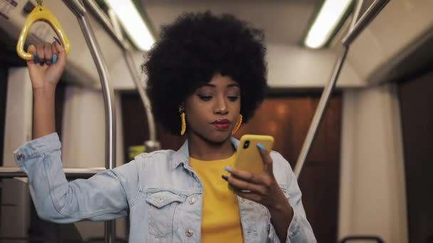 Atractiva mujer afroamericana joven sostiene la barandilla y el uso de teléfonos inteligentes en el transporte público. Noche. Primer plano. Ciudad luces fondo . — Vídeo de stock