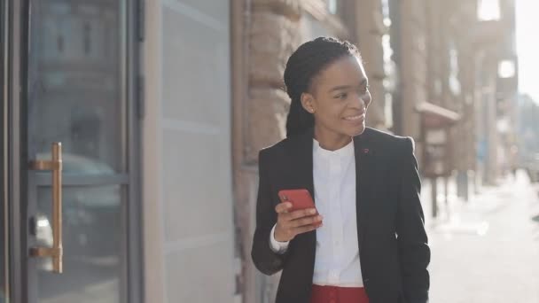 Улыбающаяся привлекательная чернокожая деловая женщина, использующая смартфон для прогулок по центру города. Черный Стиль. Дредлоки. Афро-хайрстиль . — стоковое видео