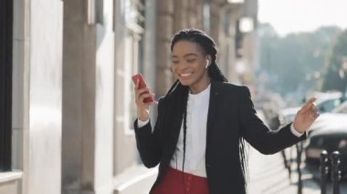 Kulaklıklı genç mutlu Afro-Amerikan iş kadını, akıllı telefondaki müziği dinleyip dans ederken şehir caddesinde yürüyor. Açık.