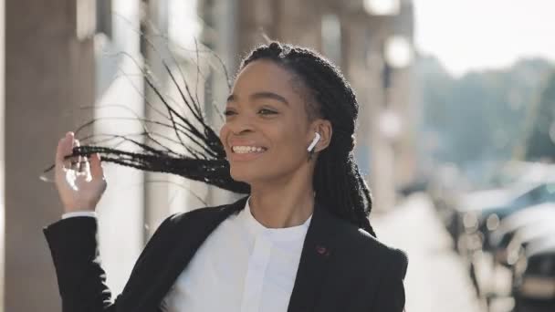 Junge fröhliche afrikanisch-amerikanische Geschäftsfrau mit Kopfhörern, die durch die Straßen der Stadt läuft, während sie Musik auf dem Smartphone hört und tanzt. Außenbereich. — Stockvideo