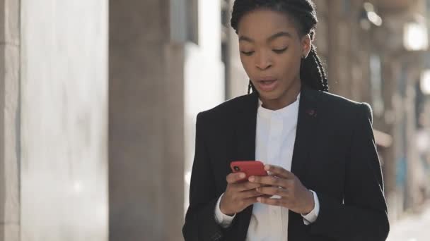 Attraktiv affärskvinna med smartphone gå på gatan nära Business Center. Svart snygg. Dreadlocks. Kvinnlig professionell i kostym ha goda nyheter på smartphone. — Stockvideo