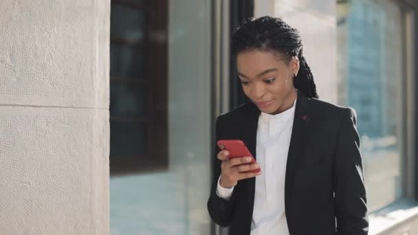 Eine elegante Afro-Geschäftsfrau steht mit ihrem Smartphone auf der Straße in der Nähe des Geschäftszentrums. schwarz stilvoll. Dreadlocks. Berufstätige haben gute Nachrichten auf dem Smartphone. — Stockvideo