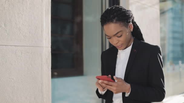 Şık afro iş kadını iş merkezi yakınında sokakta duran akıllı telefon kullanarak. Siyah Şık. Dreadlocks. Takım elbiseli kadın profesyonel akıllı telefonda iyi haberler. — Stok video