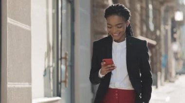 Şehir merkezinde yürüyen akıllı telefon kullanarak gülümseyen çekici siyah iş kadını. Siyah Şık. Dreadlocks. Afro Saç Modeli.