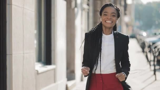 Junge fröhliche afrikanisch-amerikanische Geschäftsfrau mit Kopfhörern, die durch die Straßen der Stadt läuft, während sie Musik auf dem Smartphone hört, lustige Tänze. und schaut in die Kamera. Außenbereich. — Stockvideo