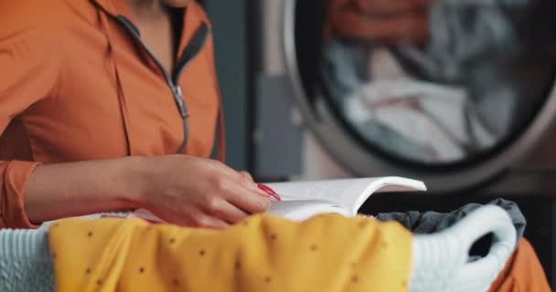 Attraktive junge afrikanisch-amerikanische Frau liest ein Buch, während sie ihre Wäsche im Waschsalon wäscht. — Stockvideo