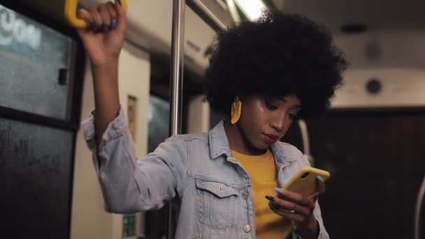 Vonzó fiatal afro-amerikai nő rendelkezik a kapaszkodó és smartphone használata a tömegközlekedés. Éjjeli idő. — Stock videók