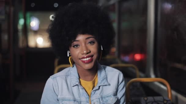 Mulher afro-americana olhando para a câmera e sorrindo sentado no transporte público, steadicam tiro. Close-up. Cidade luzes fundo . — Vídeo de Stock