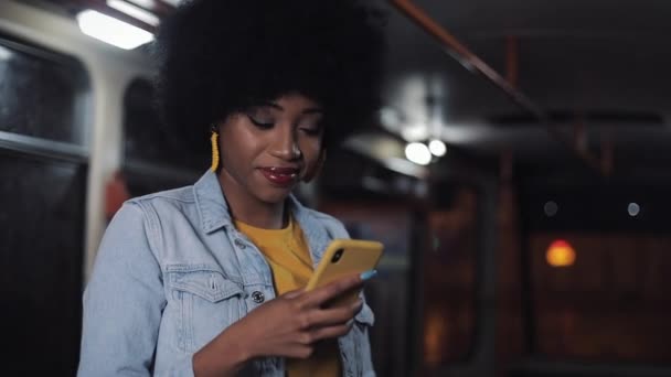 Atractiva s, iling joven afroamericana mujer usando teléfono inteligente en el transporte público. Noche. Ciudad luces fondo . — Vídeo de stock