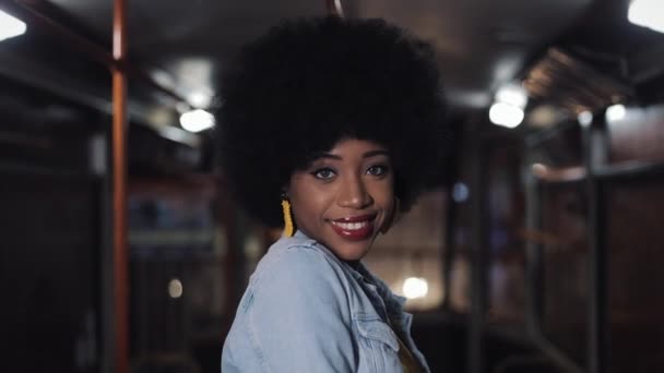 Mujer afroamericana mirando a la cámara y sonriendo de pie en el transporte público, steadicam disparó. Primer plano. Ciudad luces fondo . — Vídeos de Stock