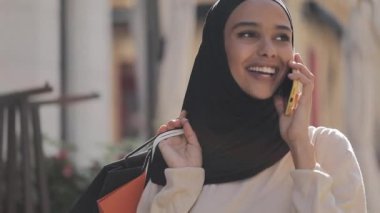 Elle alışveriş çantaları ile sokakta yürürken akıllı telefonda konuşan hijab mutlu genç Müslüman kadın.