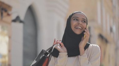 Elle alışveriş çantaları ile sokakta yürürken akıllı telefonda konuşan hijab mutlu genç Müslüman kadın.