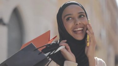 Elle alışveriş çantaları ile sokakta yürürken akıllı telefonda konuşan hijab mutlu genç Müslüman kadın.