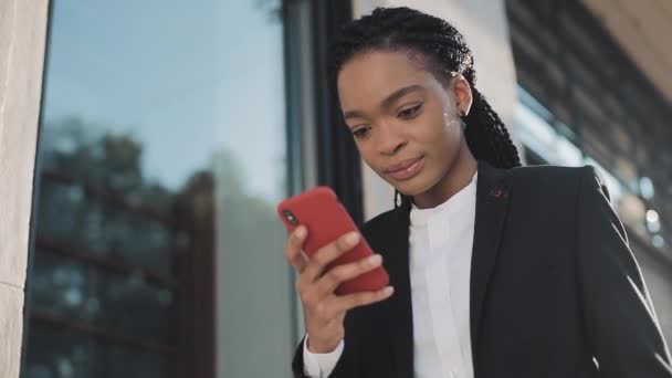 Elegante mujer de negocios afro usando smartphone parado en la calle cerca del centro de negocios. Negro con estilo. Ricitos. Peinado afro . — Vídeos de Stock