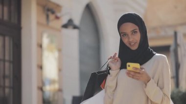 Elinde alışveriş çantaları ile sokakta akıllı telefon ile ayakta hijab mutlu genç Müslüman kadın portre. Online alışveriş, alışverişkolik kavramı,