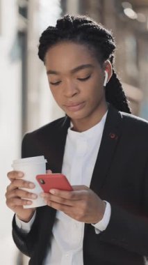 Dikey video. Eski sokak arka planda akıllı telefon kullanarak bir takım elbise kahve fincan ile Genç çekici Afrikalı Amerikalı iş kadını. Mobil cihazlar veya sosyal ağlar için dikey çerçeve.