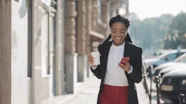 Feliz mujer de negocios afroamericana escuchando música en auriculares en el teléfono inteligente, caminando fuera de la oficina y bailando divertido al ritmo. Beber café, divertidos movimientos de baile . — Vídeo de stock