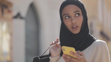 Hicap genç Müslüman kadın akıllı telefon satış arıyor, elinde alışveriş çantaları ile sokakta ayakta Internet alışveriş konsepti.