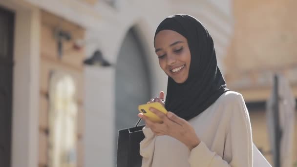 Wanita muslim muda dalam hijab mencari penjualan di smartphone, berdiri di jalan dengan tas belanja di tangannya konsep belanja internet . — Stok Video