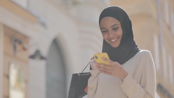 Mladá muslimka v hidžábu hledá prodej v smartphone, stojí na ulici s nákupními taškami v dlani internetové nákupy.