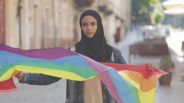 Eski sokak arka planda duran LGBT bayrağı ile başörtüsü takan güzel genç Müslüman kadın portresi. Lezbiyenlik ve İslam kavramı.
