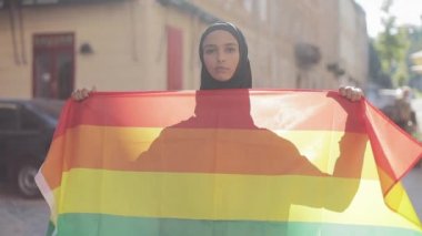 Eski sokak arka planda duran LGBT bayrağı ile başörtüsü takan güzel genç Müslüman kadın portresi. Lezbiyenlik ve İslam kavramı.