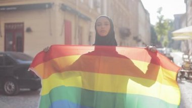 Eski sokak arka planda duran LGBT bayrağı ile başörtüsü takan güzel genç Müslüman kadın portresi. Lezbiyenlik ve İslam kavramı.