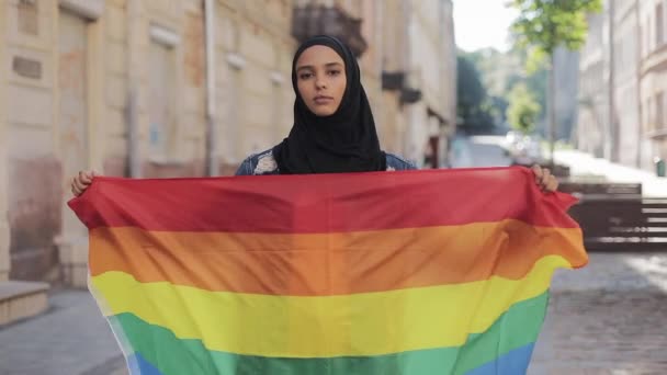 Retrato de una hermosa joven musulmana con pañuelo para la cabeza de hijab con bandera LGBT de pie sobre el fondo de la calle. Lesbianismo y concepto islámico . — Vídeo de stock