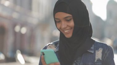 Genç gülümseyen Müslüman kadın şehir merkezinde hijab başörtüsü ayakta giyen ve akıllı telefon kullanarak. İletişim, online alışveriş, sosyal ağ konsepti.