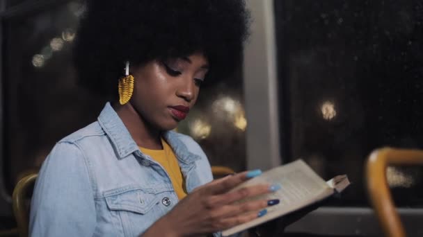 Young african american woman or passenger reading book sitting in public transport, steadicam shot. Slow motion. City lights background. — Stock Video