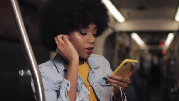 Portrait de jeune femme afro-américaine avec écouteurs écoutant de la musique, chantant et dansant drôle dans les transports publics. Il tient la main courante. — Video