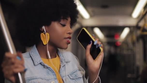Πορτρέτο της νεαρής Αφροαμερικανής γυναίκας με ακουστικά που ακούει μουσική, τραγουδούν και αστείο χορό στα μέσα μαζικής μεταφοράς. Κρατάει το τιμόνι.. — Αρχείο Βίντεο