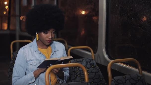 Joven afroamericana mujer o pasajero lectura libro sentado en el transporte público, steadicam disparo. En cámara lenta. Ciudad luces fondo . — Vídeo de stock