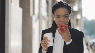 Bir takım elbise Kadın genç Afrikalı Amerikalı iş kadını bir yaz günü bir şehir caddesinde yürüyüş kahve içme online bir akıllı telefon ses tanıma fonksiyonu kullanarak.