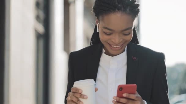 Retrato de una joven mujer de negocios afroamericana en traje, caminando por la ciudad, tomando café y usando un smartphone. Concepto: nuevo negocio, comunicación, jornada laboral, freelancer . — Vídeo de stock